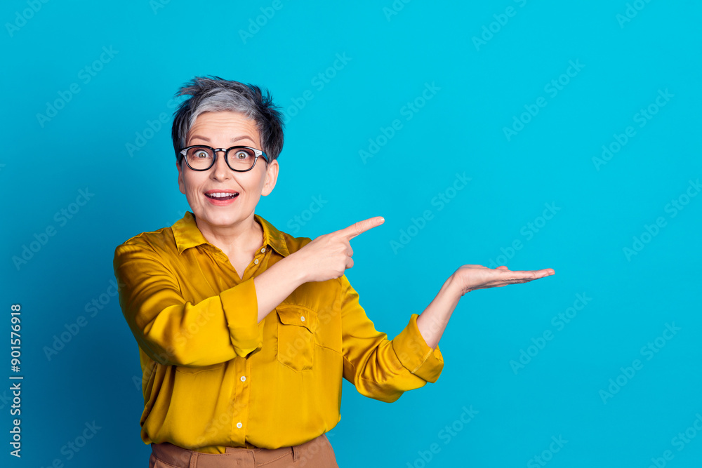 Photo of pretty cheerful woman wear blouse spectacles showing finger holding arm emtpy space isolated blue color background Photo of pretty cheerful woman wear blouse spectacles showing finger holding arm emtpy space isolated blue color background