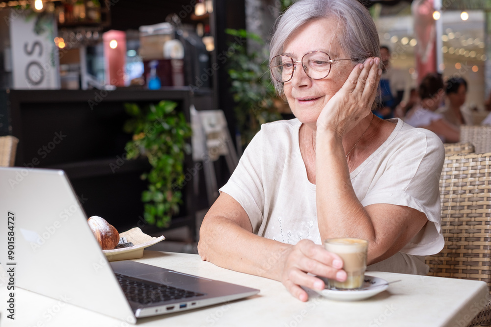 Attractive senior woman sitting in a cafe using laptop while enjoying a breakfast with pastry and coffee. Modern and smiling elderly woman surfing the net in a relaxed atmosphere Attractive senior woman sitting in a cafe using laptop while enjoying a breakfast with pastry and coffee. Modern and smiling elderly woman surfing the net in a relaxed atmosphere