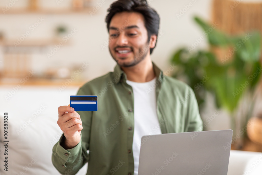 Arab man is sitting on a couch at home, holding a credit card in his right hand. He is smiling and looking at the card while using his laptop. Arab man is sitting on a couch at home, holding a credit card in his right hand. He is smiling and looking at the card while using his laptop.