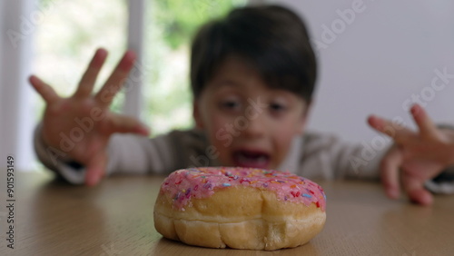 Fotografie Young boy at a table, reaching out eagerly towards a colorful sprinkled donut, d