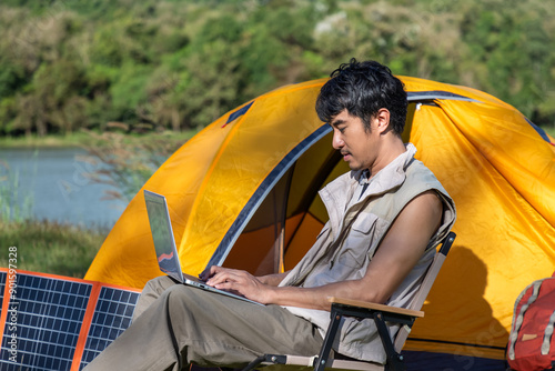 Young Asian man male sitting and remote working while camping with laptop and solar panel in an outdoor nature