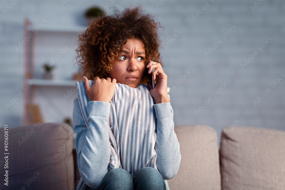 Scared afro woman calling phone and embracing pillow on sofa, afraid of noise near house, free space Scared afro woman calling phone and embracing pillow on sofa, afraid of noise near house, free space