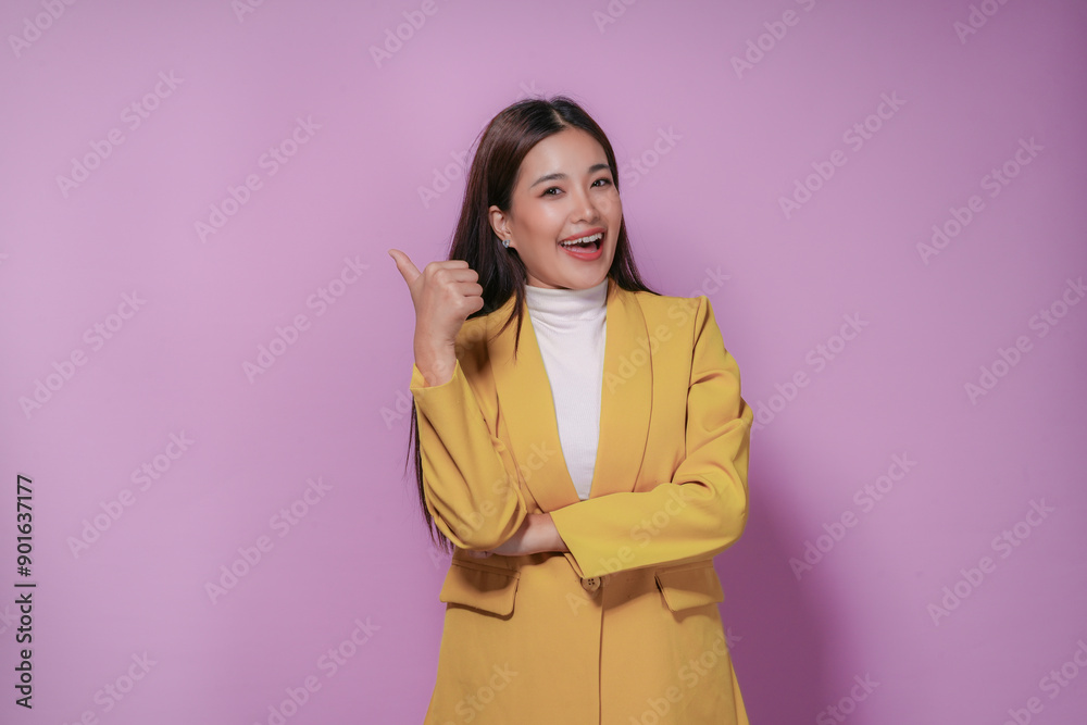 Cheerful asian businesswoman in a yellow suit is pointing her thumb to the left while smiling and looking confident on a purple background Cheerful asian businesswoman in a yellow suit is pointing her thumb to the left while smiling and looking confident on a purple background