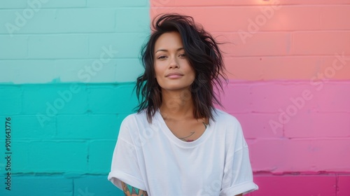 A woman with a relaxed demeanor and slightly tousled dark hair stands against a vividly colorful wall mural, exuding confidence and nonchalance in her white t-shirt. photo