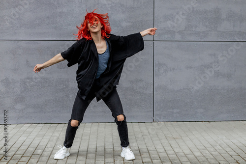 Happy positive emotional dancer curling in dance. Woman with red hair dancing on urban street, on gray wall background. International dance day. photo