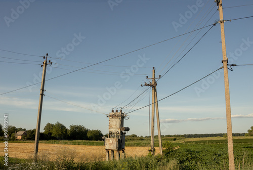 A power transformer connected to three concrete pillars around. There is rural land in the summer