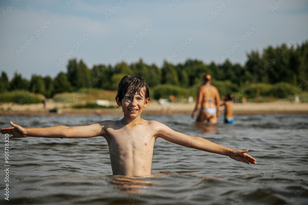 A smiling boy with arms outstretched stands in the water at a beach. Other beachgoers are visible in the background under a clear sky A smiling boy with arms outstretched stands in the water at a beach. Other beachgoers are visible in the background under a clear sky