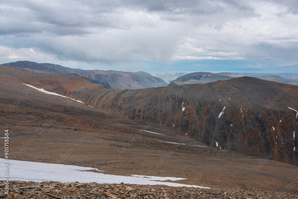 Dramatic top view from pass above large sharp ridge to alpine deep valley among big cliffs far away in rain under cloudy sky. Bleak atmospheric landscape with high rocky mountains under rainy clouds. Dramatic top view from pass above large sharp ridge to alpine deep valley among big cliffs far away in rain under cloudy sky. Bleak atmospheric landscape with high rocky mountains under rainy clouds.