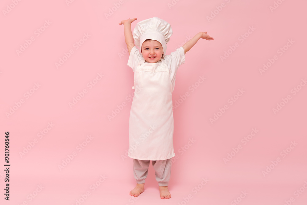 Child dressed as a chef in white uniform with arms raised. Studio portrait on pink background. Cooking and culinary concept for poster and card design. Full-length photography.