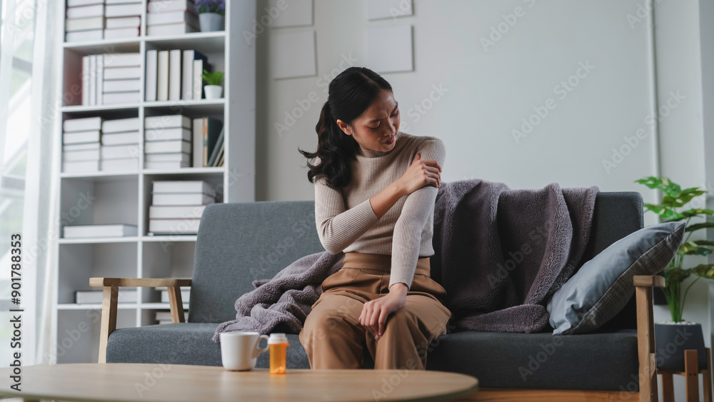 Young woman is experiencing a painful shoulder while sitting on her couch at home with a bottle of pills and a mug on the table in front of her Young woman is experiencing a painful shoulder while sitting on her couch at home with a bottle of pills and a mug on the table in front of her