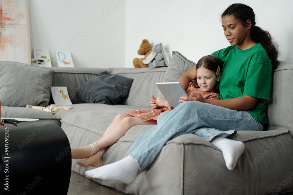 Two happy intercultural girls in casual attire watching online cartoons or curious video while both sitting on comfortable couch at home Two happy intercultural girls in casual attire watching online cartoons or curious video while both sitting on comfortable couch at home