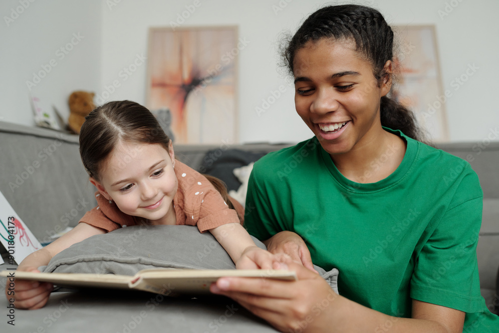 Happy teenage female sitter looking at page of open book and reading it to smiling adorable little girl pointing at text or picture Happy teenage female sitter looking at page of open book and reading it to smiling adorable little girl pointing at text or picture