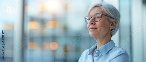 Confident senior businesswoman with short gray hair and glasses, standing in an office with a cityscape view. She is looking forward with determination, reflecting a strong and professional demeanor