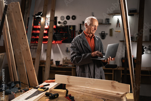 Male Woodworker Using Laptop Computer. male Carpenter Working in a Studio with a lot of Tools on the Walls. © mtrlin