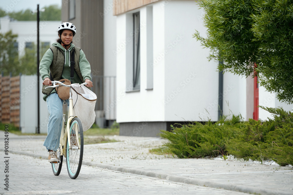 Fototapeta premium Cute African American teenager in casualwear and protective helmet sitting on bicycle with white sack containing newspapers and other post