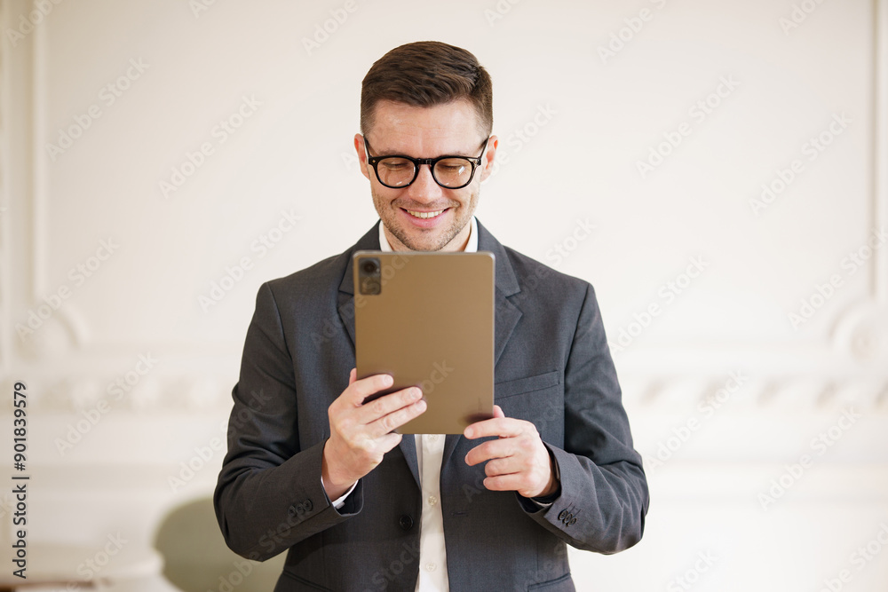 A smiling businessman in a suit and glasses reads on his tablet, standing in a bright office. A smiling businessman in a suit and glasses reads on his tablet, standing in a bright office.