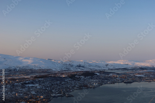 Snowy Norwegian landscape at dusk, with a distant town's twinkling lights nestled among the mountains, casting a peaceful glow and highlighting the serene beauty of this winter wonderland.