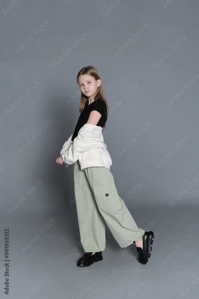 A young girl with long blonde hair, wearing a black top and light green pants, stands sideways while looking at the camera with a calm expression. The photo captures a moment of quiet contemplation A young girl with long blonde hair, wearing a black top and light green pants, stands sideways while looking at the camera with a calm expression. The photo captures a moment of quiet contemplation