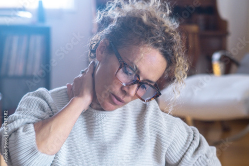One modern woman tired massaging neck and shoulder for overwork and bad posture sitting on the floor using laptop computer at home. Painful expression from neckache female at home sitting on the floor