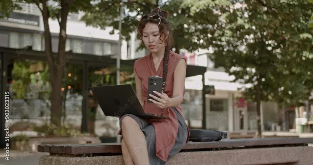 Young woman working remotely with a laptop and smart phone while sitting on a bench in an urban park. Freelance and remote work outdoors. Young woman working remotely with a laptop and smart phone while sitting on a bench in an urban park. Freelance and remote work outdoors.