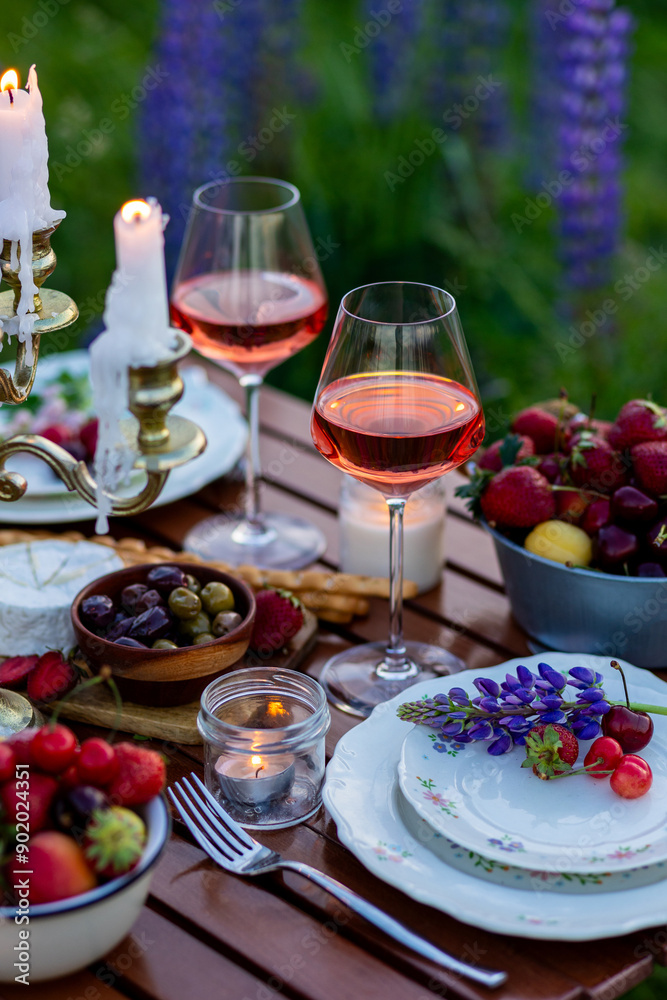 Two glasses of rose wine, flowers in a vase, silverware, fruits. Sunset, golden hour, atmospheric candlelight. Romantic table decor for a loving couple on blooming meadow with purple lupines Two glasses of rose wine, flowers in a vase, silverware, fruits. Sunset, golden hour, atmospheric candlelight. Romantic table decor for a loving couple on blooming meadow with purple lupines