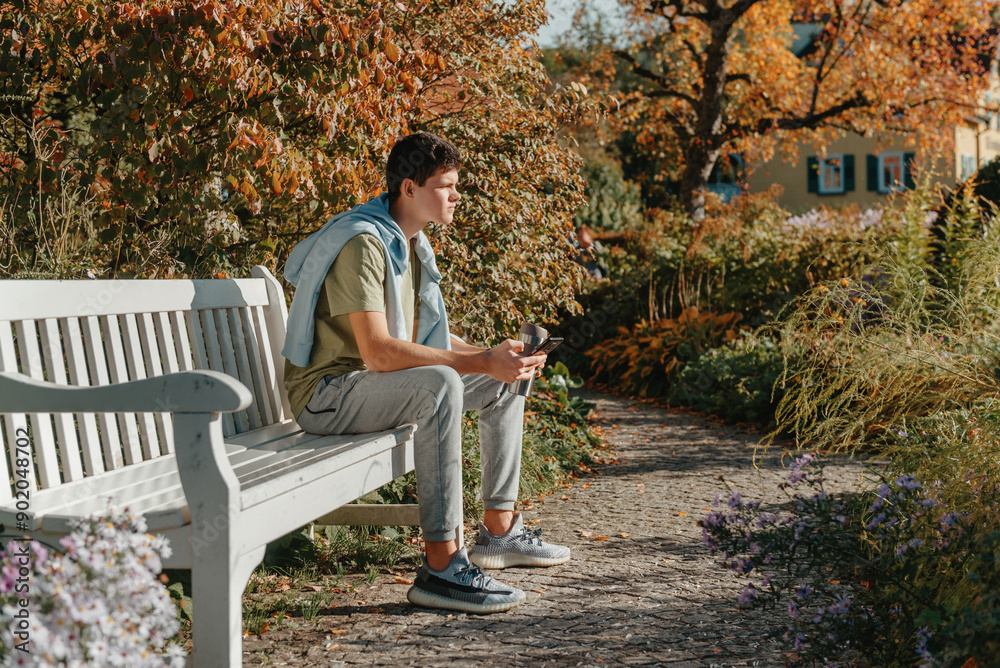 Fototapeta premium A Teenager Sits On A Bench In The Autumn Park Drinks Coffee From A Thermo Mug And Looks Into A Phone. Portrait Of Handsome Cheerful Guy Sitting On Bench Fresh Air Drinking Latte 