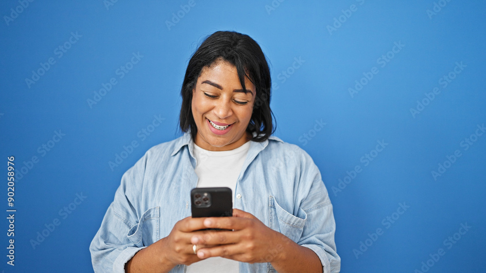 A smiling hispanic woman looks at her phone against an isolated blue wall. A smiling hispanic woman looks at her phone against an isolated blue wall.