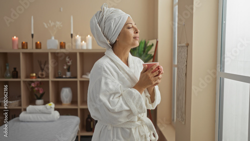 Middle-aged woman relaxing in a spa room, wearing a bathrobe and towel on her head, holding a cup, with candles and plants in the background photo