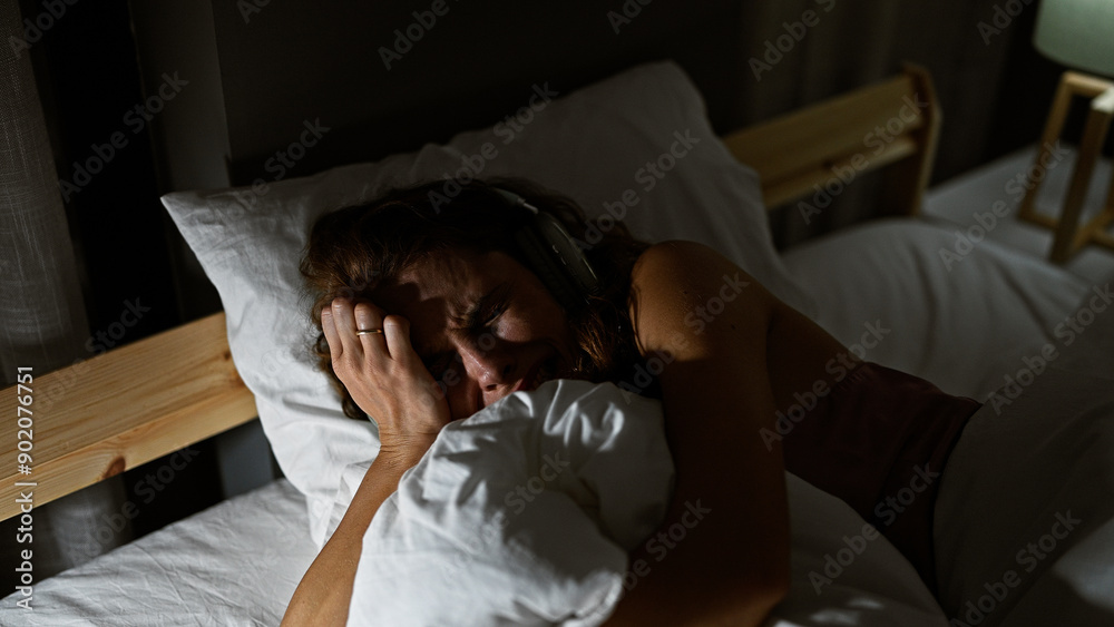 A distressed young woman in bed with headphones struggling with insomnia in a dark bedroom environment.
