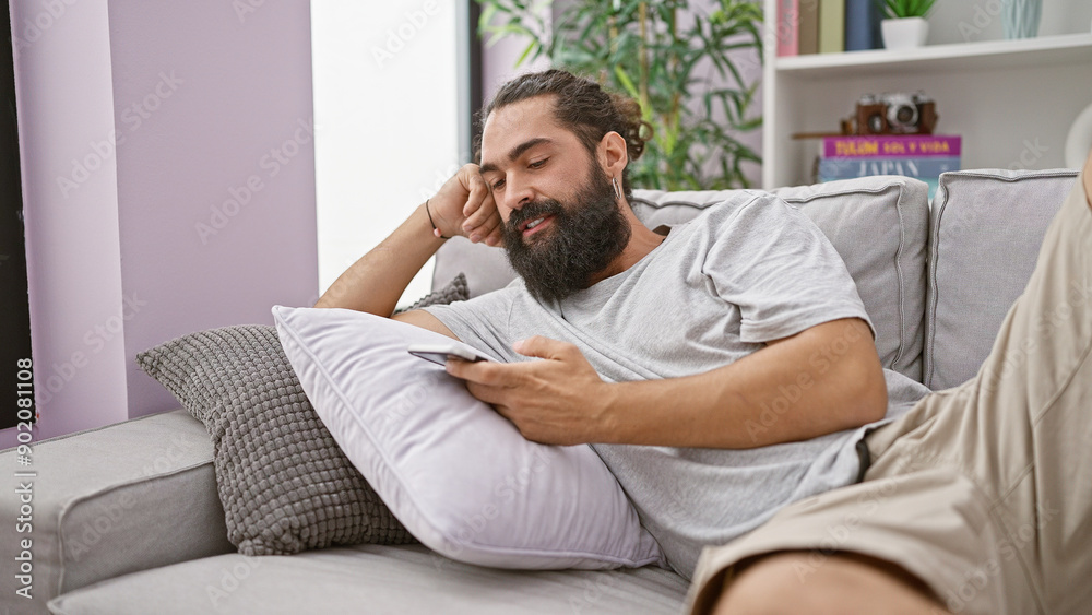 Handsome bearded hispanic man relaxing on a couch with a smartphone in a cozy modern living room Handsome bearded hispanic man relaxing on a couch with a smartphone in a cozy modern living room