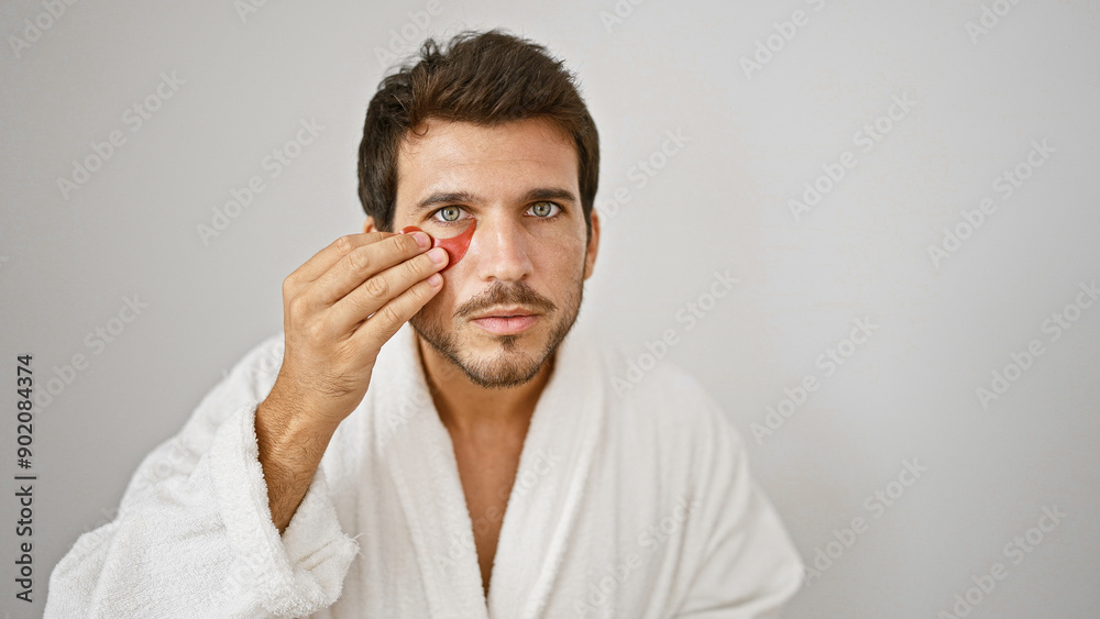 Young hispanic man in white robe applying facial skincare product against isolated white background Young hispanic man in white robe applying facial skincare product against isolated white background