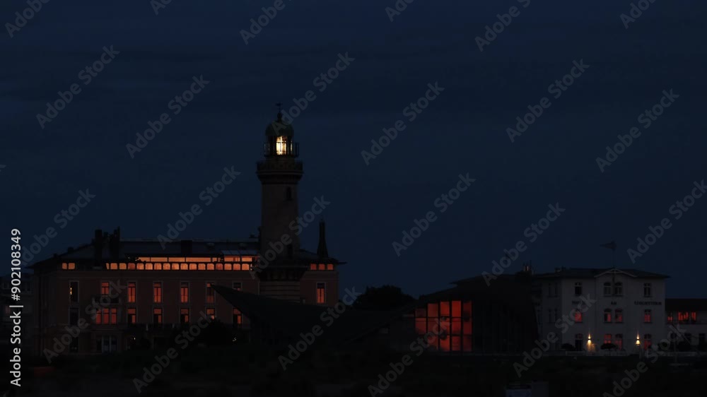 White rhythmic light beams of the famous lighthouse tower of Warnemünde, Germany in evening time. Buildings near the coast of the Baltic Sea with beautiful orange sunset reflections on their windows. White rhythmic light beams of the famous lighthouse tower of Warnemünde, Germany in evening time. Buildings near the coast of the Baltic Sea with beautiful orange sunset reflections on their windows.