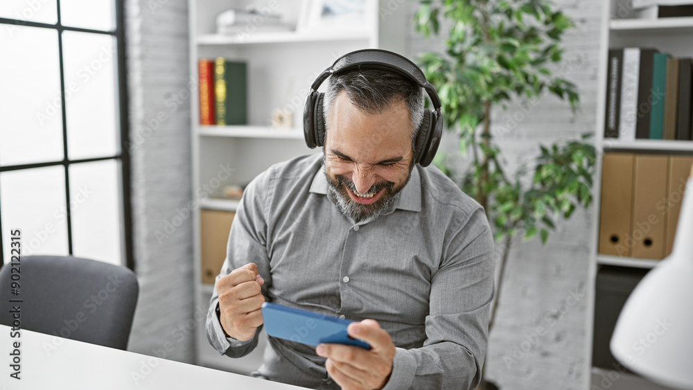 Fototapeta premium A cheerful bearded senior man with headphones plays a video game in a modern office, showcasing excitement and leisure during a break.