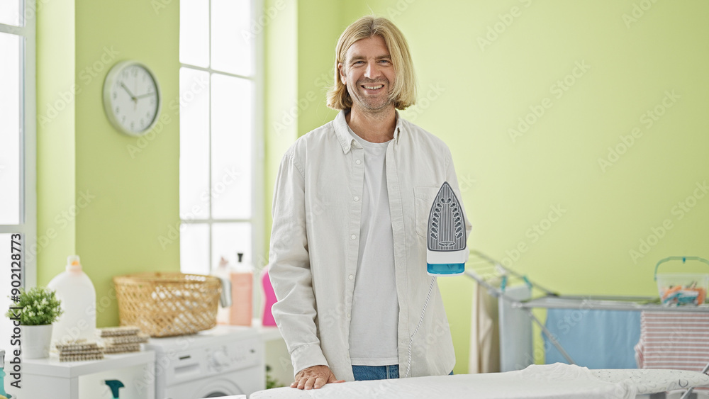 A smiling man with long blond hair ironing clothes in a green laundry room with a washing machine and laundry baskets. A smiling man with long blond hair ironing clothes in a green laundry room with a washing machine and laundry baskets.