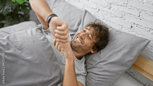 Handsome man with green eyes and beard smiling while checking time on his watch, lying in bed at a cozy home interior.