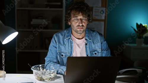 Fotografie A young hispanic man works late at an office, depicting dedication with a somber expression under desk lamp light