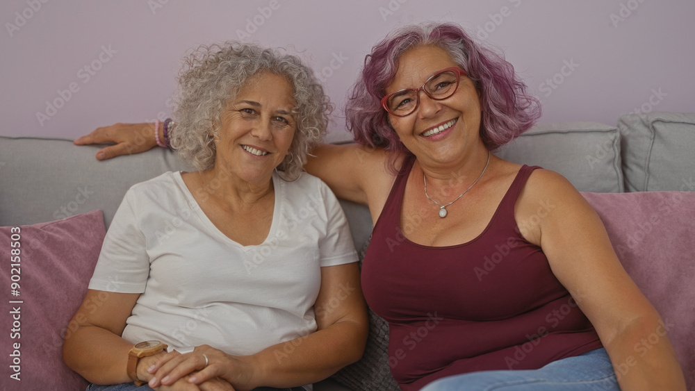 Mature women friends smiling and relaxing on a cozy sofa in a living room at home, showcasing their gray and purple hairstyles in a warm indoor setting. Mature women friends smiling and relaxing on a cozy sofa in a living room at home, showcasing their gray and purple hairstyles in a warm indoor setting.