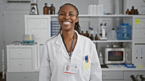 A smiling african american woman clinician with a badge stands confidently in a well-equipped laboratory room.