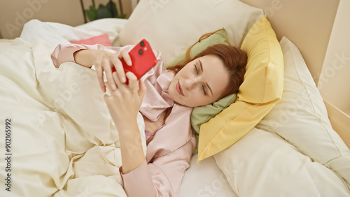 A relaxed young woman enjoys using her smartphone while lying in bed, captured in a cozy bedroom setting.