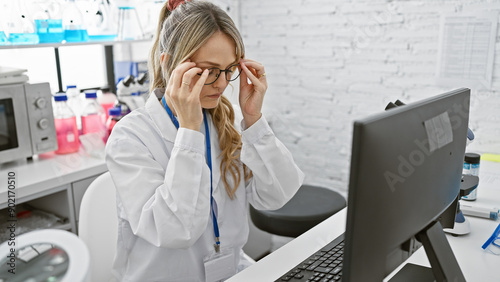 A focused blonde woman adjusts her glasses while working at a computer in a laboratory setting. photo