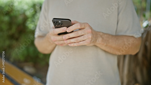 Close-up of a caucasian mature man casually dressed holding a smartphone in a calm, leafy outdoor setting.