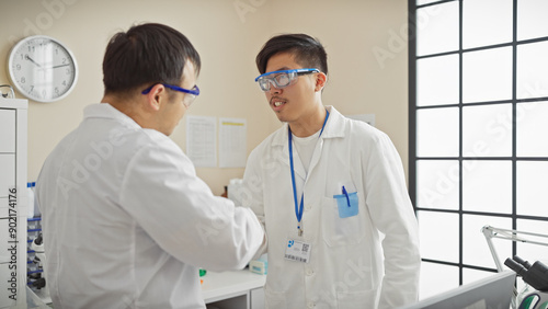 Two men in a laboratory setting discuss work, with lab coats, glasses, and id badges displayed prominently. photo