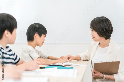 塾・学童で勉強する小学生の男の子と勉強を教えるアジア人女性講師・先生・学童指導員  © buritora