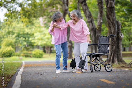 Two Elderly Women Walking Together in a Park, One Assisting the Other Near a Wheelchair, Demonstrating Care, Support, and Friendship on a Bright Day © kamonrat