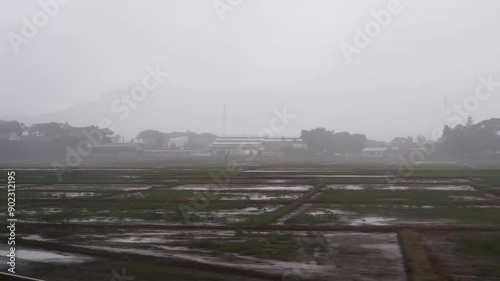 Wide shot sideways POV, small farm fields and houses in the rain and fog from a speeding train, out of Kendal, Central Java Indonesia