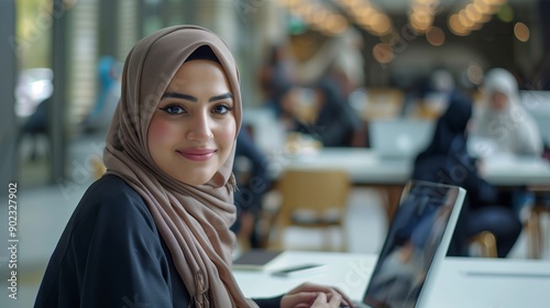 A young Muslim woman in hijab works at her laptop and smiles while sitting at the table