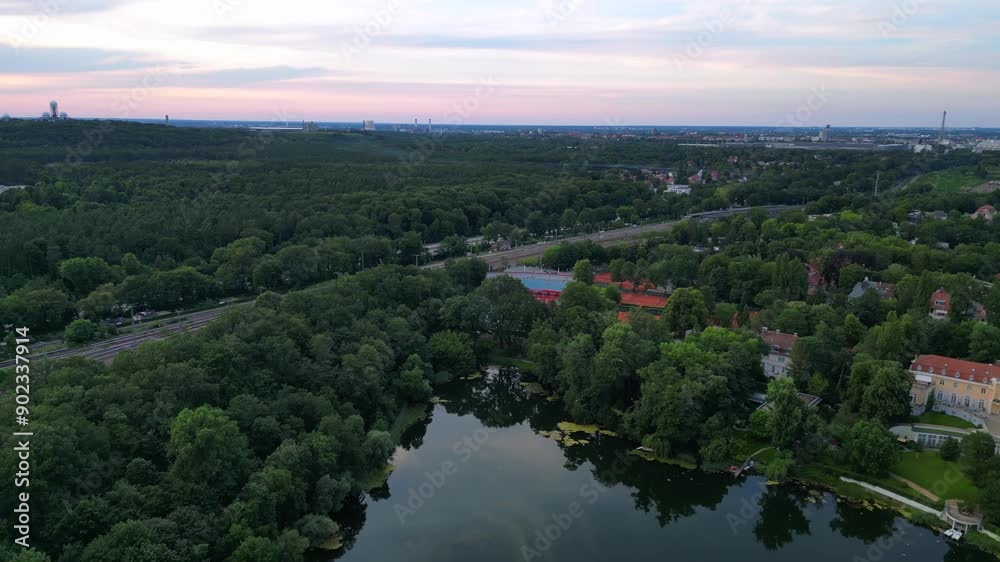 Tennis stadium with red clay courts, surrounded by a lush green forest and a small lake. Marvelous aerial view flight descending drone