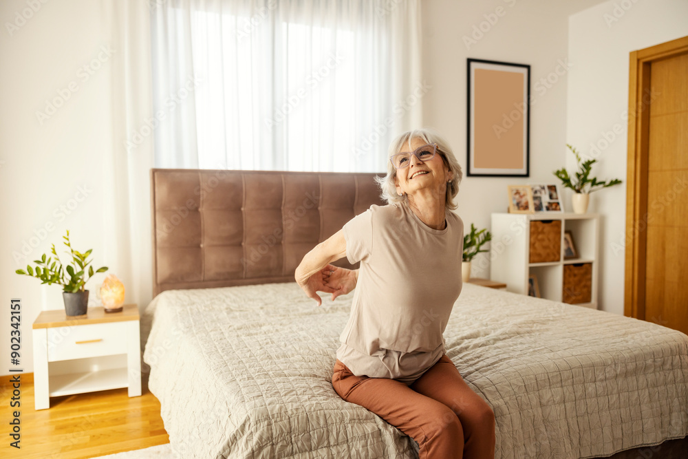 Happy senior woman is stretching her arms in the morning while sitting on a bed in bedroom. Happy senior woman is stretching her arms in the morning while sitting on a bed in bedroom.