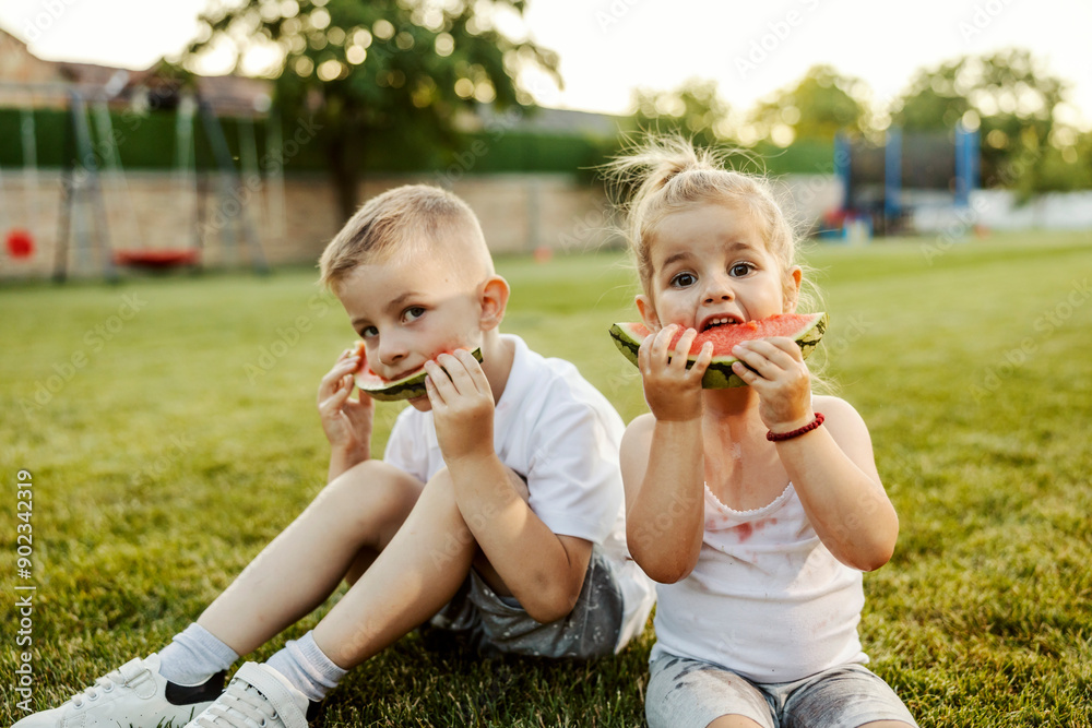 Adorable little brother and sister sitting on a lawn in backyard and eating juicy watermelon. Adorable little brother and sister sitting on a lawn in backyard and eating juicy watermelon.