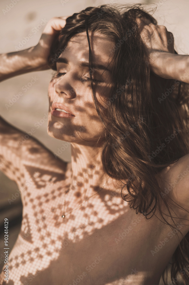 Fototapeta premium Portrait of a beautiful woman with long hair in abstract shadows on the background of sand in the desert. Fashion art shooting in gold color with lace and shadows on the face